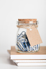 paper clips in glass jar with blank paper tag on stack of notepads. Office working space. mockup for branding. copy space. office stationery concept