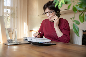 Beautiful stylish elderly woman talking on a mobile phone while working at home with a laptop.