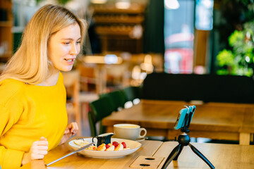 Blond female blogger holding fork and knife. Woman in modern cafe interior eating cottage cheese pancakes or traditional syrniki with with raspberries jam with yogurt. Healthy breakfast concept.
