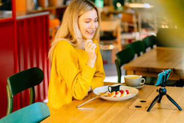 Caucasian woman is happy to communicate with foreign friends or family via smart phone on tripod in modern interior of coffee shop.Blogger communicates with subscribers via video link during breakfast