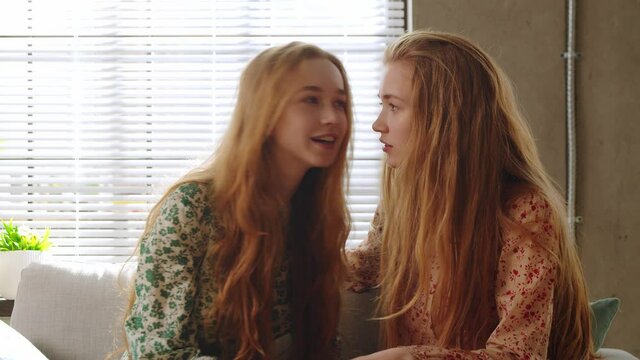 Beautiful twin sisters have fun talking, sharing secrets and laughing sitting on the sofa in a cozy living room at home