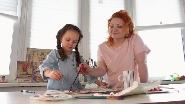 Family art leisure activity concept. Mother and daughter painting together at home using paintbrushes and watercolors. Attractive woman standing and helping or teaching daughter to draw. 4k footage