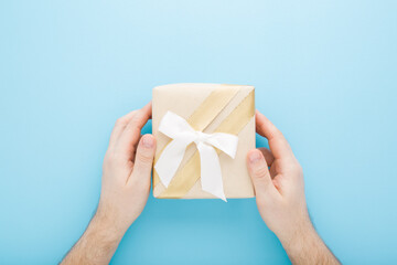 Young adult man hands holding beige paper gift box with white ribbon on light blue table background. Pastel color. Closeup. Point of view shot. Congratulation concept. Top down view.