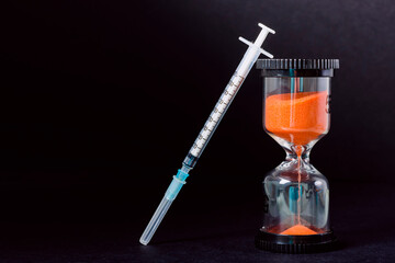 Hourglass and syringe on black background.The photograph is a horizontal shot and aims to create...