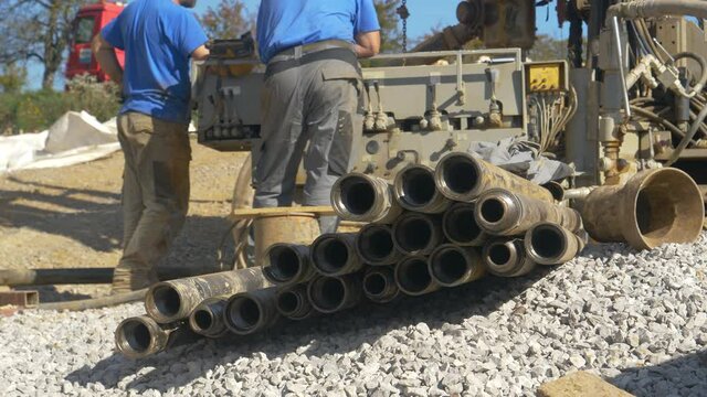 LOW ANGLE, CLOSE UP, DOF: Thick metal pipes lie on the gravel ground near a big boring rig. Detailed shot of pipes as contractors operate a massive drill excavating soil near modern housing project