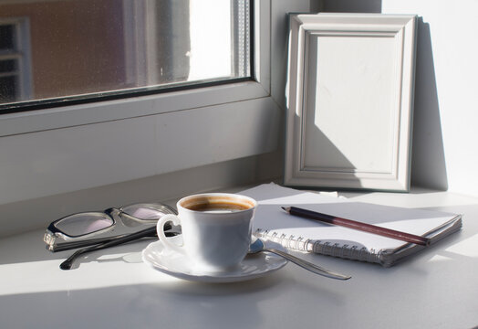 A white porcelain cup with strong coffee in a saucer, glasses, a notebook with a pencil, an empty picture in a frame on the windowsill against the background of the window.