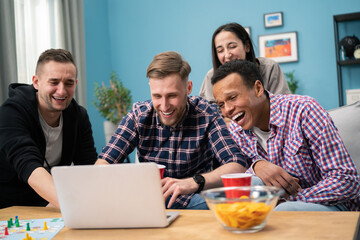 Happy diverse friends watching funny video, laughing at joke, using laptop, having fun together, sitting on couch, looking at computer screen, students colleagues working on online project