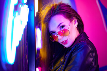 Fashion portrait of a young woman in sunglasses posing near neon signs in night club