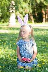 Cute little girl wearing bunny ears is sitting on a green grass and holding painted Easter eggs