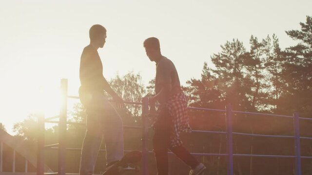 Slowmo tracking shot of young man skating up ramp and giving high-five to his friend. Then they are skating down ramp in light of setting sun