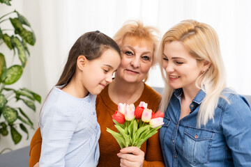 Fototapeta na wymiar A little girl gives a bouquet of tulips to her mom and grandmother. They celebrate Mother's Day. They have a traditional family holiday.