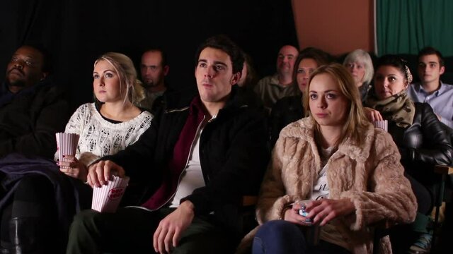 Caucasian Couple sat in Cinema Theatre watching a film or movie. He tries to put arm round her. Stock Video Clip Footage