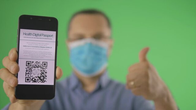 Young man with face mask encourages to use the qr-code scanable display of digital health certificate vaccination passport for COVID-10 coronavirus, thumb up front view isolated on green screen chroma