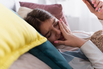 Fototapeta na wymiar frustrated teenage girl covering mouth while crying and holding smartphone in bedroom
