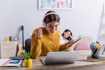 happy teenage girl in wireless headphones holding notebook and pen near laptop