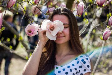 A lovely young woman with dark hair holds a magnolia flower near her face. White magnolia in the botanical garden. Girl with flowers in a white dress with polka dots.