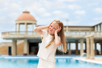 Fototapeta premium Emotions. Portrait of a little girl holding her head and showing her displeasure. Headache. In the background there is a building with a dome and a swimming pool