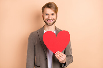 Photo of cheerful sweet young guy wear jacket smiling holding large red heart isolated beige color background