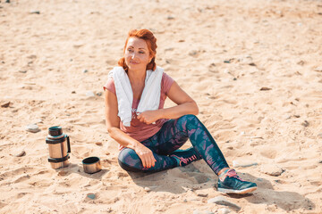 After training. Smiling adult woman in sports clothes, sitting on the sand. Next to it is a bottle of sports nutrition. The concept of sports lifestyle and sports nutrition