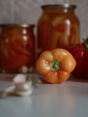 Vertical close up shot of an orange bell pepper and a garlic with jars of canned bell peppers in the backdrop. 