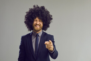 Happy cheerful toothy smiling adult caucasian businessman bearded wearing curly black toupee and elegant formal suit pointing index finger towards camera studio portrait over grey background