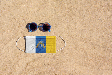A medical mask in the color of the Canary Islands flag lies on the sandy beach next to the glasses. The concept of organizing a safe beach holiday in Canary Islands during the coronavirus pandemic.