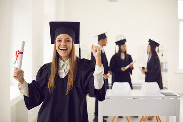 Super happy, triumphant, excited graduate standing in university or college room, holding diploma and clenching fist. Smiling student proud of academic achievements celebrating her graduation - obrazy, fototapety, plakaty