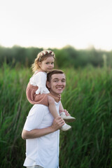 cute young dad with his daughter toddler, baby girl sitting on dad's neck, walking in the park in summer. Fatherhood concept. Soft focus and toning