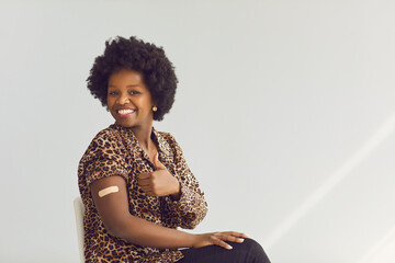 Vaccinated young smiling african american woman with medical patch on hand showing thumbs up gesture approving Covid-19 vaccination studio portrait. Antiviral immunization promotion concept - obrazy, fototapety, plakaty