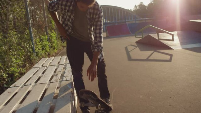 Slowmo handheld tracking shot of young man riding skateboard and sitting down on pallets in skatepark, then taking his mobile phone out of pocket and browsing internet