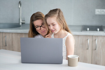 Mother with teen daughter sitting on the modern kitchen and watching a video on a gray laptop. Home Education concept