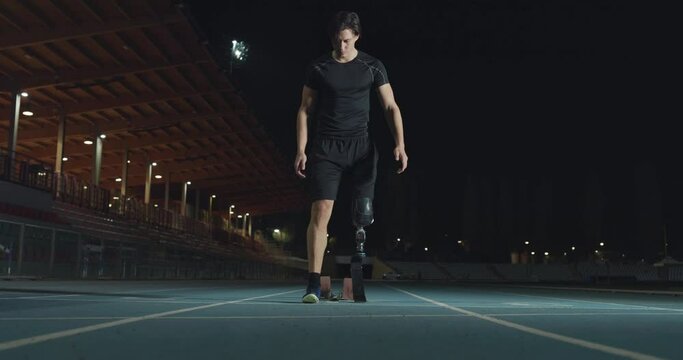 Cinematic shot of young disabled man with legs prosthesis prepares for launching off the start line on race track at night. Concept of handicapped people active lifestyle, determination.