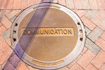 Grungy manhole cover that says Communication and USA in brick sidewalk with diagonal shadow - top down view.