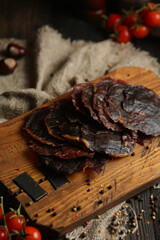Delicious smoked sausage on a dark wooden board with tomatoes and spices. Delicious handmade snack made from natural ingredients. Pieces of dry pork, beef, chicken snacks. Meat products. Close-up.