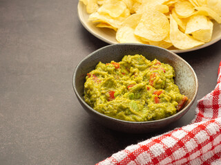 Fresh guacamole and crispy potato chips on a plate placed on dark brown background with copy space for text. Top view. Concept of traditional Mexican foods and healthy fruits