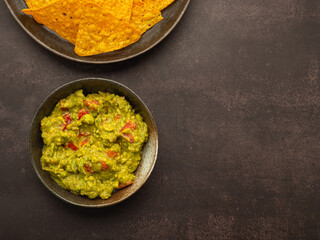 Fresh guacamole and tortilla chips on a plate placed on dark brown background with copy space for text. Top view. Concept of traditional Mexican foods and healthy fruits