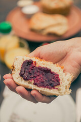 Hand holding a slice of bread with strawberry jam on the foreground. Breakfast unfocused at the background.