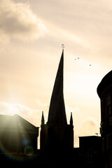 Fototapeta premium Chesterfield Crooked Spire church silhouette of tower in town centre with sun setting behind and birds in sky cockerel weather vein on top. Historic St Marys parish worship building.