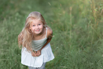 Fototapeta premium A small blonde girl of 6 years old stands among the green grass and looks mischievously at the camera