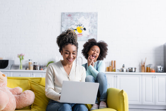 excited african american girl laughing near mother working on laptop at home