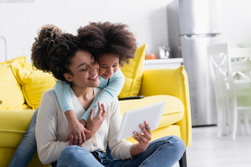 happy african american woman showing digital tablet to daughter embracing her at home