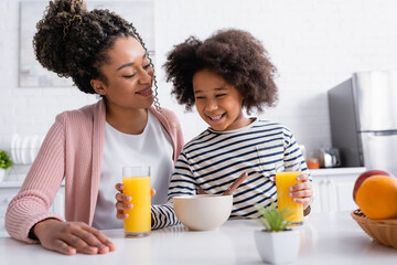 cheerful african american girl holding orange juice near mother during breakfast, blurred foreground