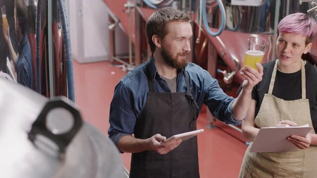 Medium PAN shot with slowmo of couple of professional brewery workers looking at glass with beer sample produced at factory, checking its color and quality