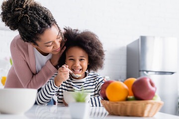 african american woman touching hair of happy kid eating corn flakes near fruits on blurred...