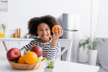 joyful african american kid holding orange near wicker basket with fruits on blurred foreground