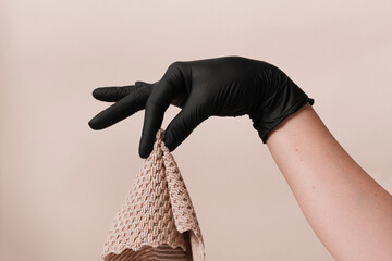 One female hand wear black disposable glove holding beige cloth above taupe background.
