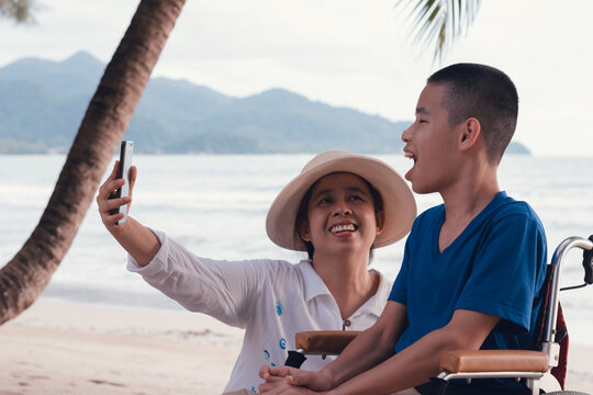 Disabled Child On Wheelchair Is Smiling,use Smartphone With Parent On The Sea Beach Like Other People,Lifestyle Of Special Child, Life In The Education Age Of Children, Happy Disability Kid Concept.