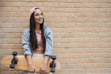 Smiling asian woman wearing stylish casual clothing holding skateboard standing near wall, copy space 