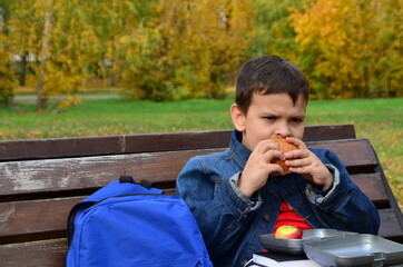 Close-up of a small schoolboy sitting on a Park bench and opening his school backpack during a lunch break. eating a sandwich out of a lunch box