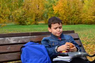 Obraz premium Close-up of a small schoolboy sitting on a Park bench and opening his school backpack during a lunch break. eating a sandwich out of a lunch box